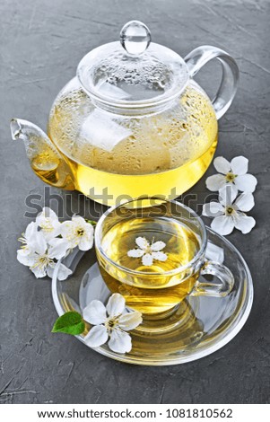 Close-up glass teapot and teacup with green tea and spring flowers on dark gray concrete background