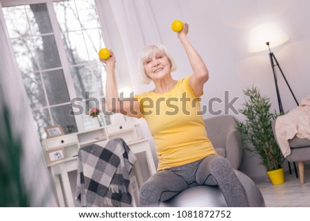 Great mood. Upbeat senior woman sitting on the yoga ball and exercising with dumbbells while smiling happily