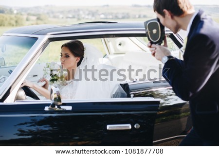 Groom making photo of beautiful bride in wedding dress with flover bouquet on the old vintage, retro camera. Bride woman sitting in luxury retro car. Wedding photoshoot