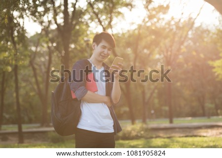 Smile asian young man hugs book while his standing use smartphone during sunset 