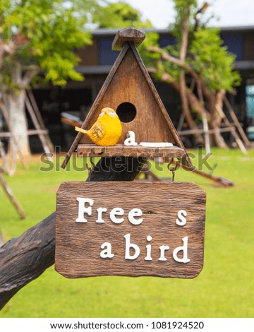 Close up of vintage wood sign with text " free as a bird" and yellow bird sculpture, bird house with gable roof decorated in garden