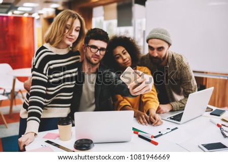 Young team of creative multicultural students making photo on front camera of modern smartphone during break in university.Casual dressed hipster guys taking selfies on mobile phone sitting at laptops