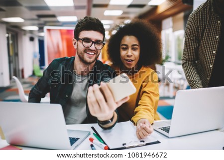 Positive carefree young man with african american student making funny selfies on smartphone sitting at laptop devices in office.Cheerful two friends taking photos on front camera of telephone