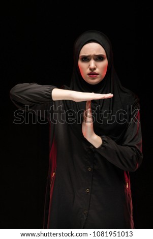 Portrait of beautiful desperate scared frightened young muslim woman wearing black hijab showing stop sign on black background