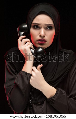 Portrait of beautiful serious scared frightened young muslim woman wearing black hijab calling for help on black background