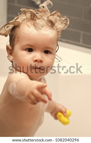 Little pretty baby boy standing in bathroom with wet foam crazy hairdo holding yellow duckling toy hold out hand, vertical picture