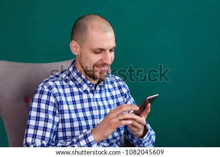 
a man with a beard and a cutoff is sitting on a gray chair against the green wall and working at the computer and talking on the phone