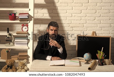 Teacher and school supplies in classroom. Alternative education and knowledge concept. Man with beard on white brick wall background. Professor with serious face sits at desk.