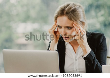Worried young attractive businesswoman sitting at table and looking at laptop screen while touching temples. Front view with big window and blurry green view outside in background.