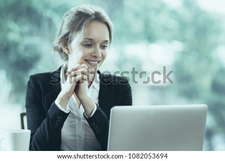 Smiling young attractive businesswoman sitting at table while looking at laptop screen with her hands clasped. Front view with big window and blurry green view outside in background.