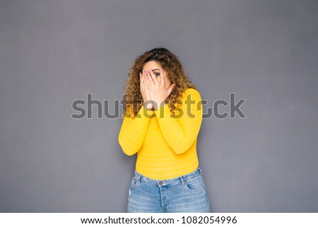 Cute brunette plus size woman with curly hair in yellow sweater and jeans standing on a neutral grey background. She scared, hides her eyes and face under her arms