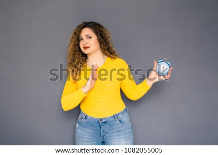 Cute brunette plus size woman with curly hair in yellow sweater and jeans standing on a neutral grey background. She holds alarm clock, unhappy what she sees, she late on her meeting or deadline