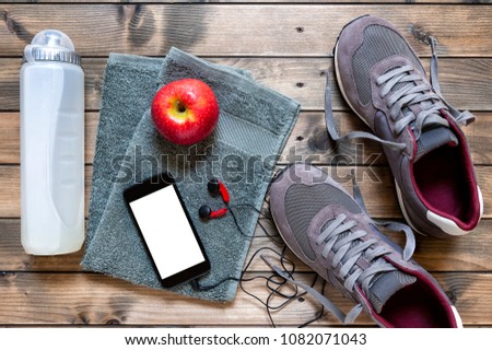 Top view of a red apple, sport shoes, audio headphone, smartphone, towel and water bottle photographed on an antique wooden table. Text space.