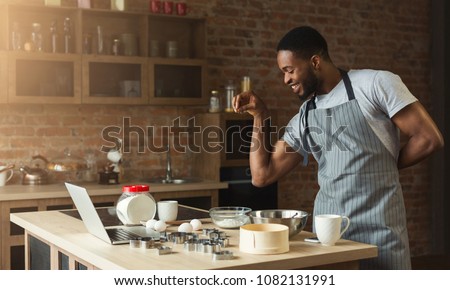 Black man seasoning food at home kitchen. African-american guy in apron baking cookies, adding flaworings to dough and having fun Royalty-Free Stock Photo #1082131991