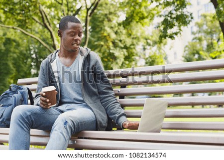 Sad handsome young african-american man using laptop outdoors, having coffee. Technology, disappointment, education and remote working concept, copy space
