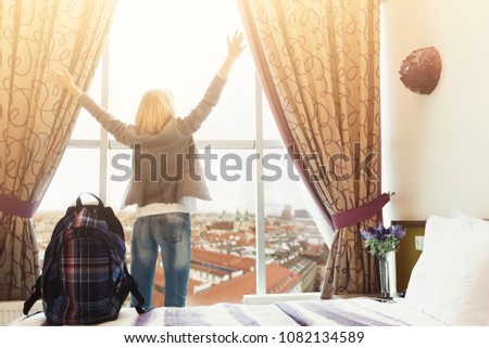 Daybreak at hotel room. Young female traveller watching cityscape, standing near big window. Happy girl with backpack ready to explore new city. Travelling and accomodation concept, copy space Royalty-Free Stock Photo #1082134589