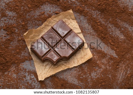 Milk chocolate bar with piece bitten off and small pieces on gray background sprinkled with cocoa powder, copy space. Confectionery shop advertising and cooking ingredients concept, mockup for recipe