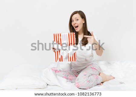 Young brunette woman sitting in bed with red striped gift box, white sheet, pillow, wrapping in blanket isolated on white background. Beauty female spending time in room. Rest relax good mood concept