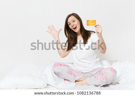 Young fun happy woman sitting in bed with white sheet, pillow, wrapping in blanket on white background. Beauty female holding credit card with money in room. Rest, relax good mood concept. Copy space