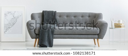 Grey couch with dark blanket standing in bright interior with metal table and simple poster Royalty-Free Stock Photo #1082138171