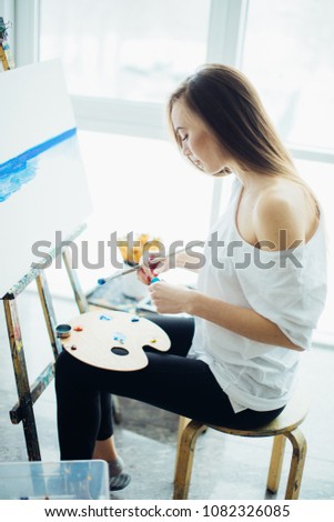 European woman painter with palette on her kneels , drawing a painting at workshop flooded with day light.