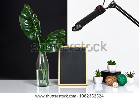 The stylish interior with mock up poster frame, leaves in glass bottle on table with black and white wall on background.