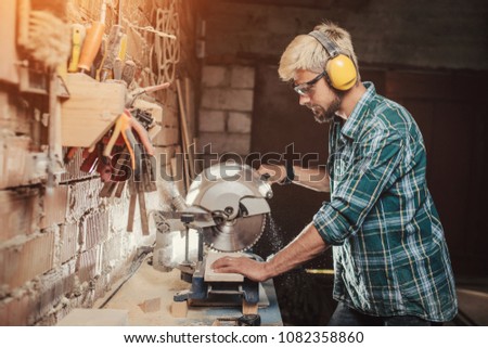 Young hipster bearded man with ear protectors by profession carpenter builder saws with a circular saw a wooden board on a wooden table in the workshop. Royalty-Free Stock Photo #1082358860