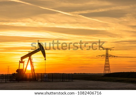 Pumpjack and transmission towers at sunset symbolizing energy transition. The setting sun is passing between the posts of a pump jack with electricity pylons and power line against a red sky.