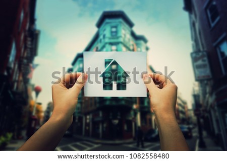 Hands holding a paper with house icon on a big city street. Find ideal place for home. Property insurance concept. Stay home social media campaign during COVID-19 quarantine isolation. Social distance