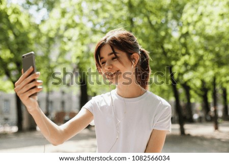 Young hipster brunette girl making a selfie or video with front camera on mobile phone, student having a funny video call and laughing in the park during break in university, wifi in public places