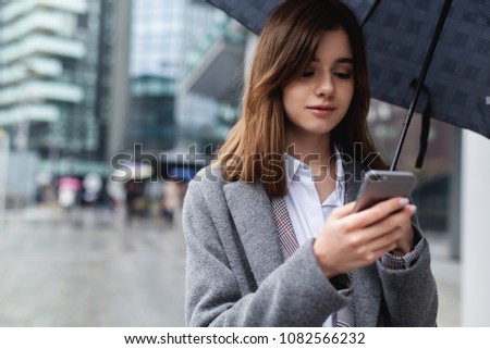 Brunette businesswoman using mobile phone near office, young girl browsing phone smiling under umbrella while it is raining outdoors, female manager texting smartphone near modern bank building