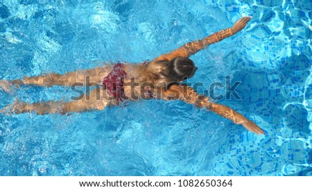 Unrecognizable beautiful girl in red bikini floating across the pool of hotel. Young woman swimming in clear blue water of basin on sunny day. Concept of summer vacation or holiday. Top view Close up.