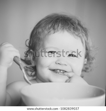 Portrait of funny little smiling boy with blonde curly hair and round cheecks eating from green plate holding spoon closeup, square picture