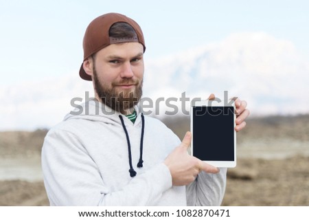 Close-up portrait of a smiling hipster in a brown cap outdoor holding a white tablet computer with a cut-out blank for the designer. A bearded man looks at the tablet. background blur.