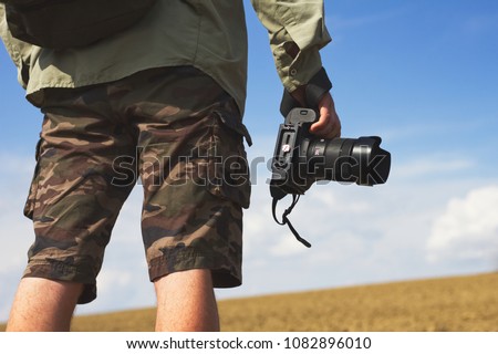 Photographer holding a camera outdoors. Man wearing camouflage shorts taking pictures of nature. 