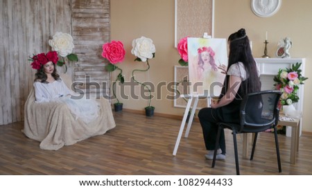 artist draws a portrait from nature. Beautiful model, with a wreath of scarlet peonies on his head, posing sitting in a white armchair