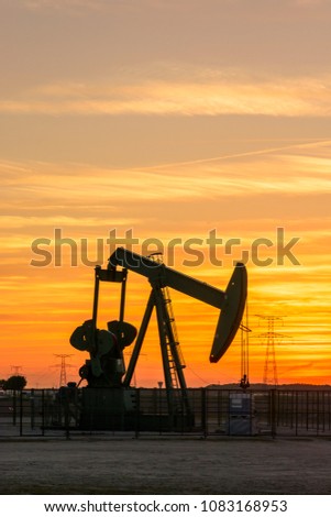 Pumpjack and transmission towers at sunset symbolizing energy transition. A pump jack pumping oil out of a well with silhouettes of electricity pylons and power line against a red sky.