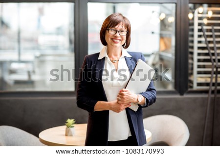 Portrait of a senior business woman dressed in the suit standing with laptop in the modern cafe interior