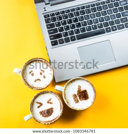 Smile emoji painted on cups of cappuccino next to the laptop on a yellow background. Emotions and communication online concept