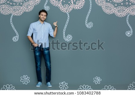 Cool and attractive. Full length of young charming man keeping right hand in pocket and pointing back with the thumb of his left hand