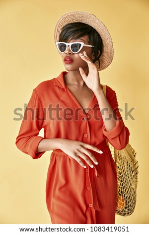 Colorful portrait of young black woman with short brown hair and big red lips, weared in red orange dress, hat, white sunglasses, with fruits in net bag. Yellow background Royalty-Free Stock Photo #1083419051