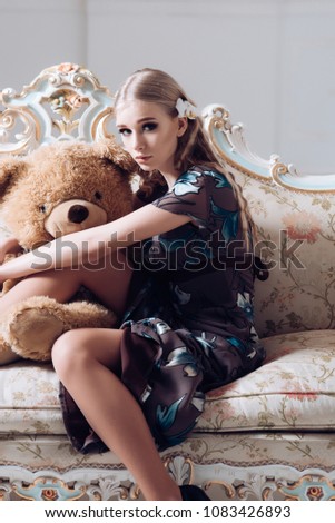 Lovely blond girl hugging huge teddy bear while sitting on retro sofa. Slim woman in flowery gray dress in her room.