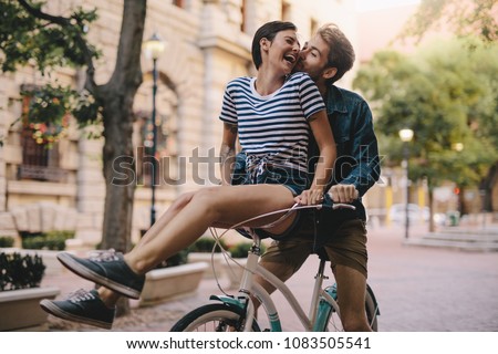 Laughing woman sitting on boyfriends bicycle handlebar. Cheerful couple on a bike together in the city having fun. Royalty-Free Stock Photo #1083505541