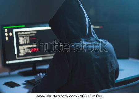 cybercrime, hacking and technology concept - male hacker in dark room writing code or using computer virus program for cyber attack Royalty-Free Stock Photo #1083511010
