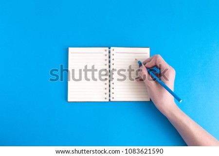 Female hand writing in blank notebook against blue background minimal creative concept. Space for copy.