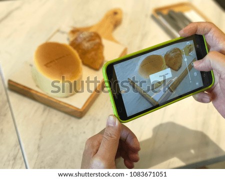 A man is taking a picture of a baked croissant and a cheese cake on the wooden plate with his smart phone.