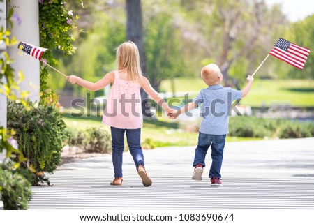 Young Sister and Brother Holding Hands and Waving American Flags At The Park.