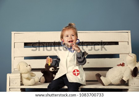 Happy baby in vet doctor uniform with toy pets sit on wooden bench on blue background. Health, healthcare, medicine. Veterinary clinic game. Future profession concept.