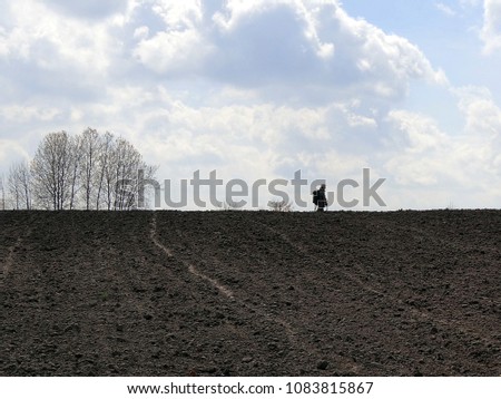 A man with a metal detector on a plowed field, against a background of sky and clouds