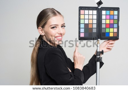 Young photographer pointing at color checker card, while working in studio. Girl in formal suit prepares for photo shooting. Photography concept. Woman on smiling face stands near color checker card.